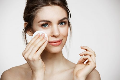 Cute beautiful natural brunette girl cleaning face with cotton sponge smiling looking at camera over white background. Cosmetology and spa. Copy space.